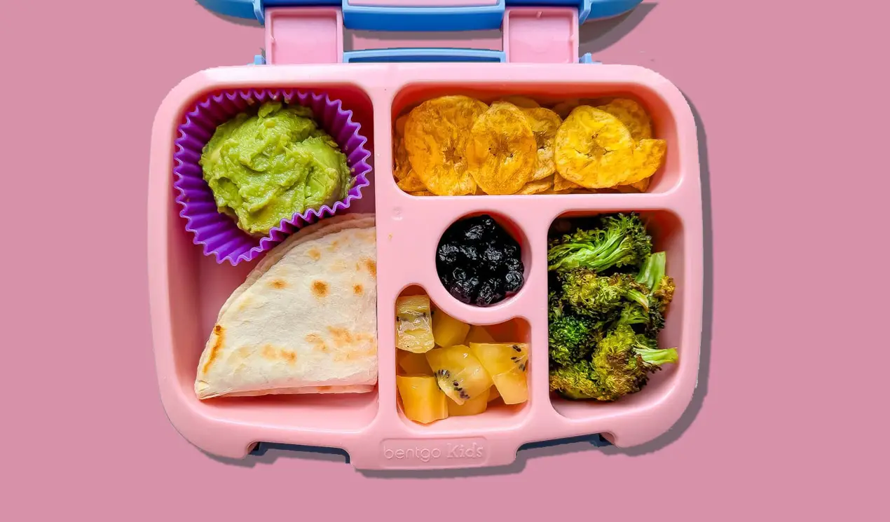 A lunch box with cheese quesadilla, guacamole, plantain chips, roasted broccoli, yellow kiwi and dried blueberries. A lunch box with cheese quesadilla, guacamole, plantain chips, roasted broccoli, yellow kiwi and dried blueberries.