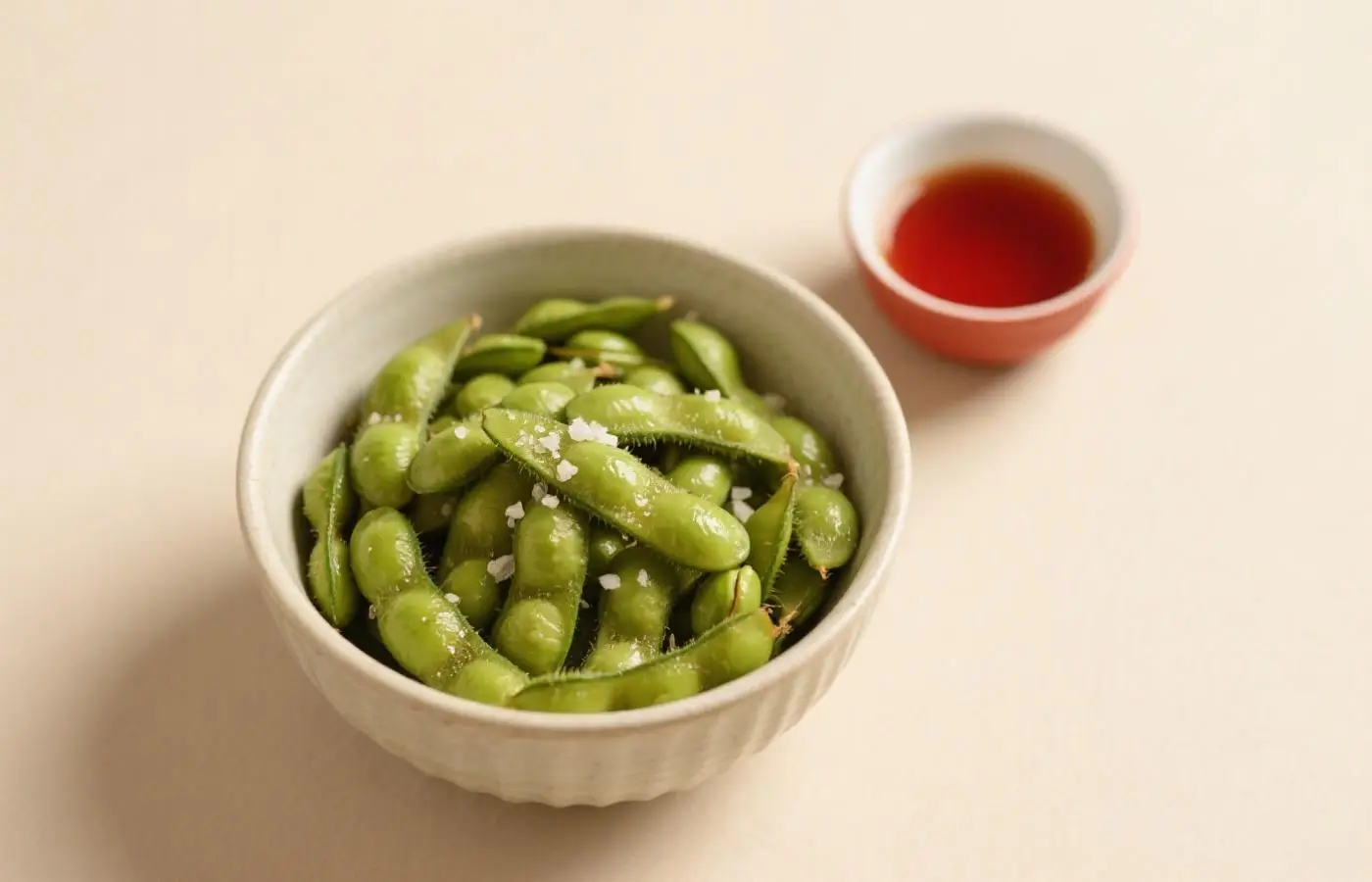 A bowl of edamame, which is yellowed green in color and sprinkled with white sea salt on top. The edo sprouts have not yet thrown away the seeds at one end. A small red sauce sits next to it on the table. The background light beige tablecloth creates an atmosphere that resembles Japanese cuisine. A bowl of edamame, which is yellowed green in color and sprinkled with white sea salt on top. The edo sprouts have not yet thrown away the seeds at one end. A small red sauce sits next to it on the table. The background light beige tablecloth creates an atmosphere that resembles Japanese cuisine.