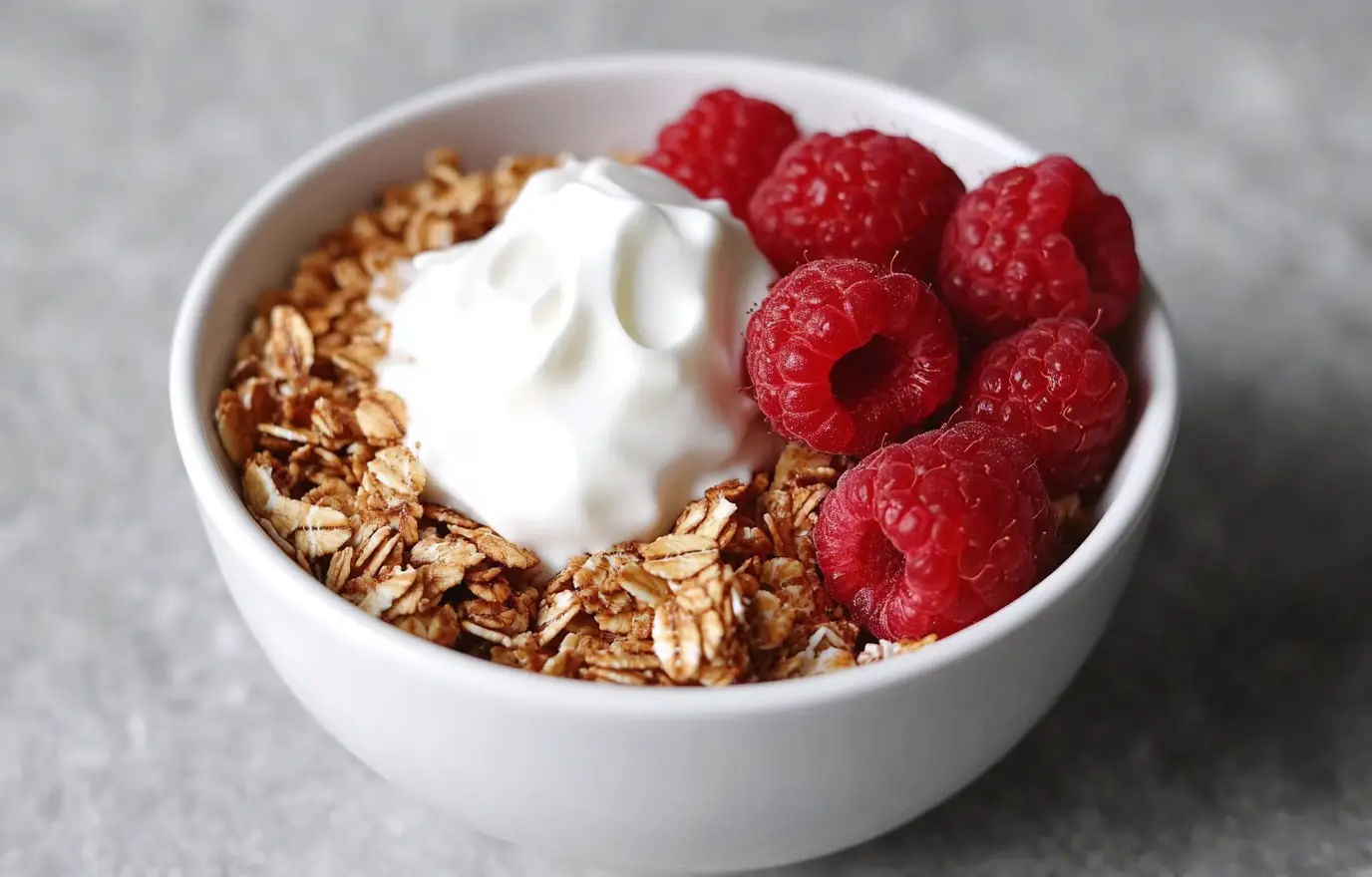 A white bowl filled with granola, red raspberries, and Greek yogurt on top of the granola, set against a beautiful light grey background. The granola is on the right side of the picture. The photo is taken from above, looking down at an angle. There are no other items around it. It looks delicious. A white bowl filled with granola, red raspberries, and Greek yogurt on top of the granola, set against a beautiful light grey background. The granola is on the right side of the picture. The photo is taken from above, looking down at an angle. There are no other items around it. It looks delicious.