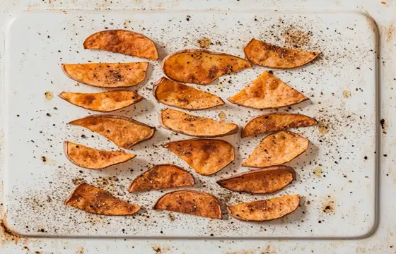 A photograph of crisp and thin sweet potato chips with black pepper powder on top, arranged neatly on an empty white baking sheet. The potatoes have a golden-brown color, and visible spices like salt or paprika are sprinkled over them. There is no text present in the photo.