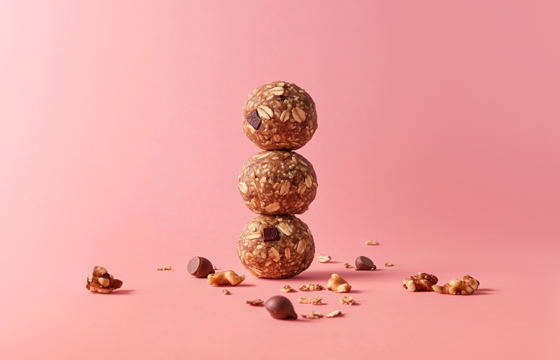 A stack of three energy balls, with one on top, sitting against a pink background. The spheres have an earthy brown color and visible chocolate chips speckled throughout the surface. Some broken granola pieces surround them. A few small oak fuzzy nuts can be seen scattered around. The scene is well-lit, with soft lighting that highlights their texture and shape.