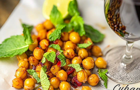 "Cuban". A photo of roasted chickpeas with chili powder, lime juice, and mint leaves. The texture is golden brown, small in size, oval-shaped, and smooth to the touch. They have an intense, zingy impact from the tropical colors of light yellow, green, red, and purple. They are placed on top of a paper kitchen towel. High-angle view, macro photography.