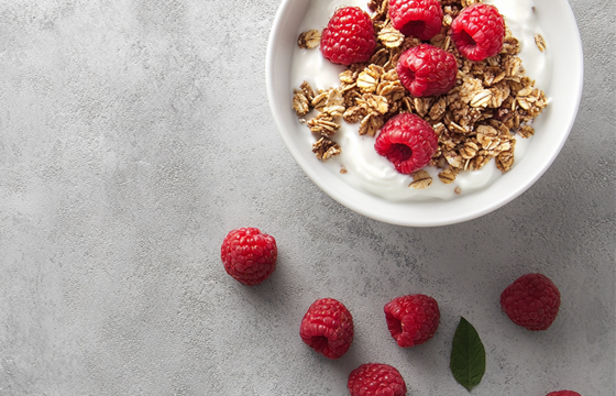 A photo of a white bowl filled with granola, topped with Greek yogurt and fresh raspberries. The background is light gray concrete, captured from above in a close-up shot. Soft natural lighting illuminates the scene, highlighting the textures and colors of each element. Taken with an iPhone camera using portrait mode for sharp focus and vibrant color.