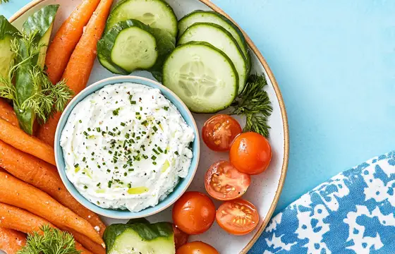 A bowl of white cream cheese and chives on the side, surrounded by carrots, tomatoes, cucumber slices, and some green leaves. The background is light blue, and there's space for text at the top right. On a plate, there was also an all-blue patterned cloth napkin under it.