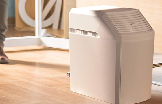 A beige whole - house humidifier placed on a wooden floor in an indoor setting, with a partial view of a person and furniture in the background