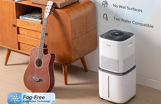 A white and gray humidifier with “Fog - Free Evaporative Technology” label, featuring “No Wet Surfaces” and “Tap Water Compatible” benefits, placed next to a wooden cabinet with a guitar on the floor.