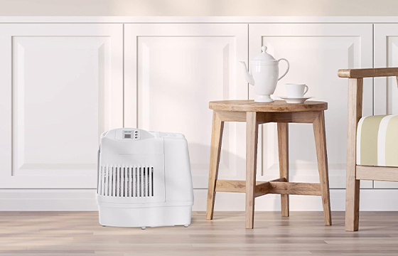 A white compact humidifier situated on a wooden floor beside a small wooden table with a white teapot and cup, against a white paneled wall.